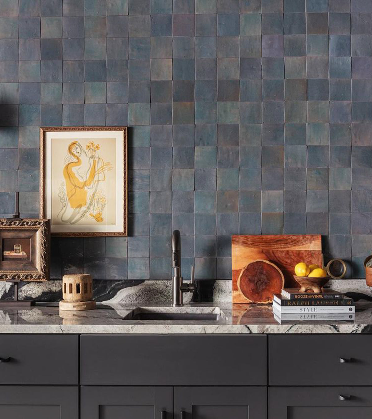 A kitchen counter with dark cabinetry, a marble countertop, stacked books, lemons, cutting boards, a framed art print, and a textured Graphite Grey 4x4 tile backsplash by vendor-unknown.