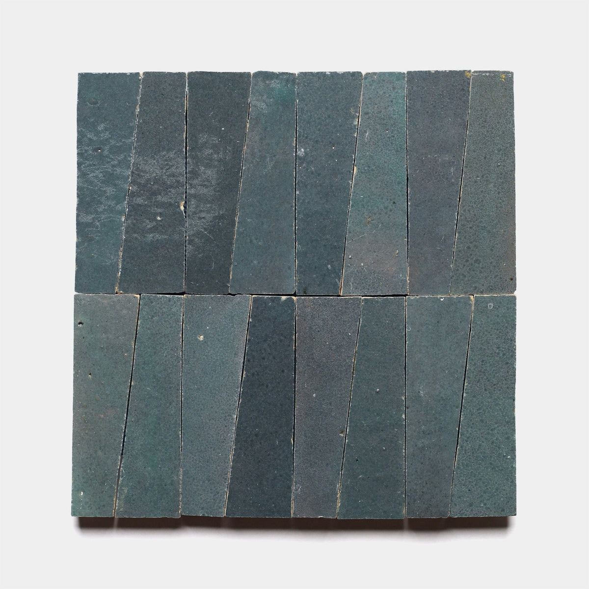 Sixteen rectangular Graphite Grey Trapezoid Sample tiles by Zia Tile, arranged in a 4x4 grid, display subtle texture variations and slightly irregular shapes in a dark greenish-blue hue.