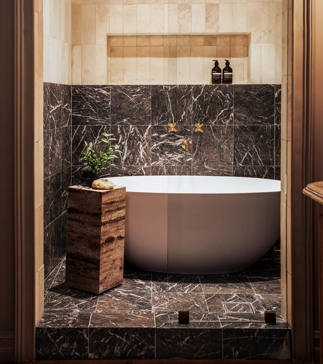 A modern bathroom features a white oval tub against Zia Tile’s Grigio Carnico 12x12 marble tiles, gold fixtures, two bottles on a shelf, and a stone table with a plant and sponge. Warm lighting adds coziness.