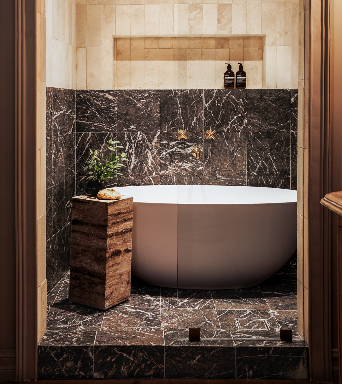 A modern bathroom features a white oval tub against Zia Tile’s Grigio Carnico 12x12 marble tiles, gold fixtures, two bottles on a shelf, and a stone table with a plant and sponge. Warm lighting adds coziness.