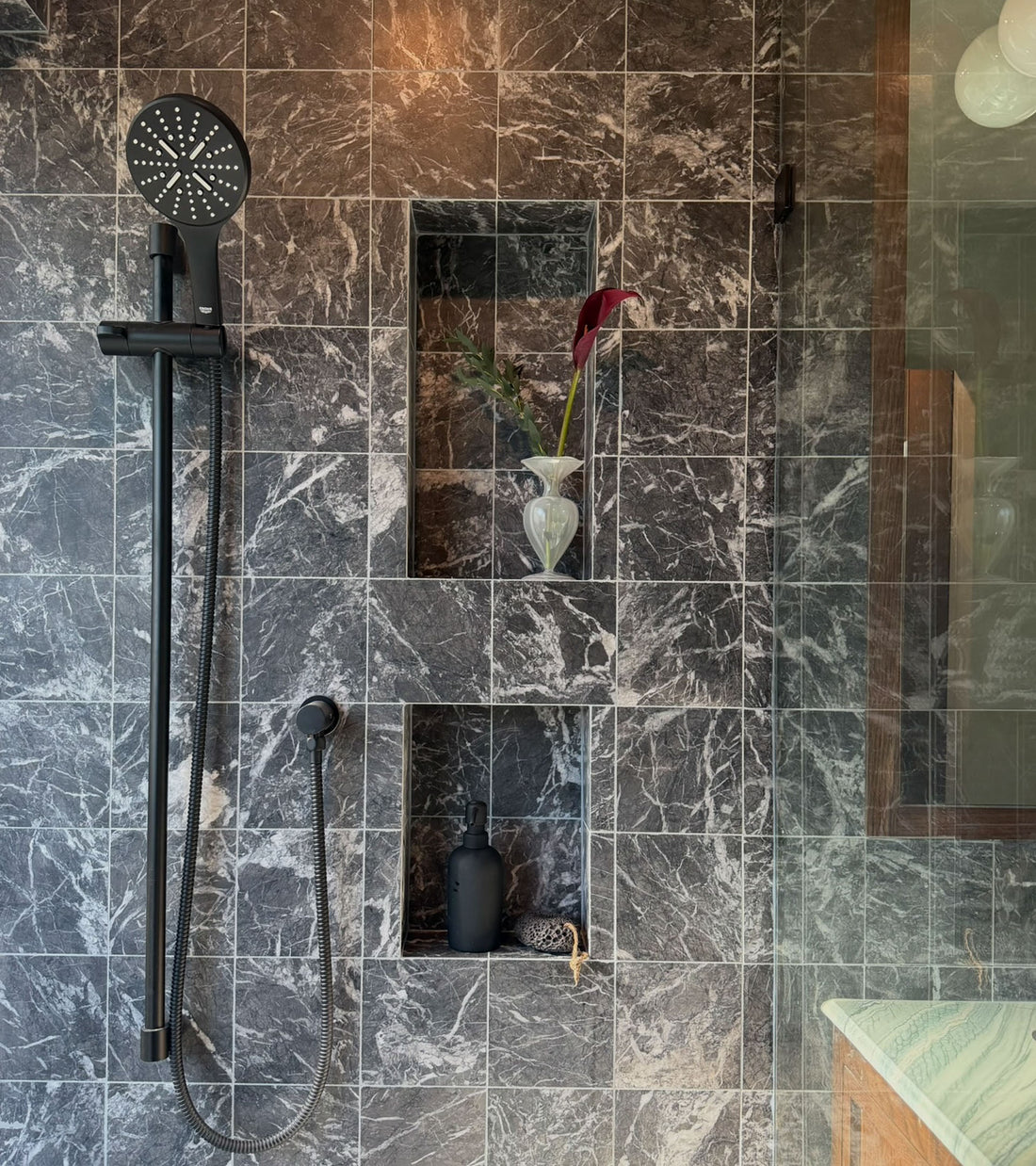 A modern shower features Zia Tile's Grigio Carnico 6x6 dark gray marble tiles, a black handheld showerhead, two wall niches with a white vase and black bottle, and part of a glass shower door.
