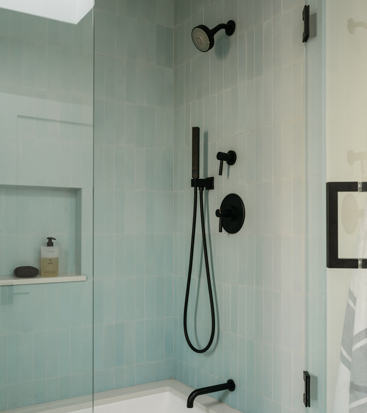 A modern shower features vendor-unknown Hailstone 2x8 blue subway tile in a vertical layout, a black showerhead, handheld sprayer, and faucet. A glass door encloses the space. Built-in shelf holds soap and lotion; nearby hook holds a towel.