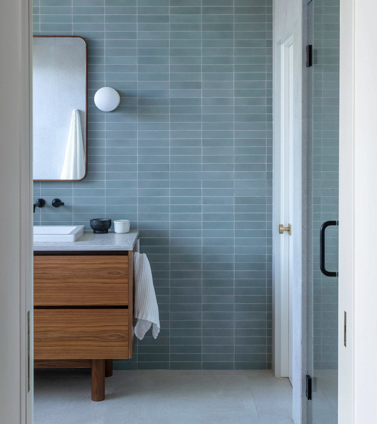Modern bathroom featuring walls with Hailstone 2x8 tiles by vendor-unknown, a wooden vanity with white sink, round white wall light, rectangular mirror, glass shower door with black handle, and a white towel on the vanity.