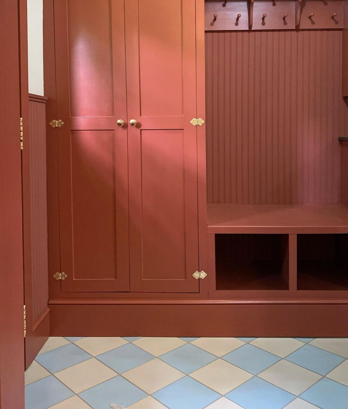 A terracotta red built-in mudroom unit with brass-knob cabinets, open cubbies, and a bench sits atop vendor-unknown Hailstone 8x8 cement tile in a blue and beige diamond pattern for added charm.