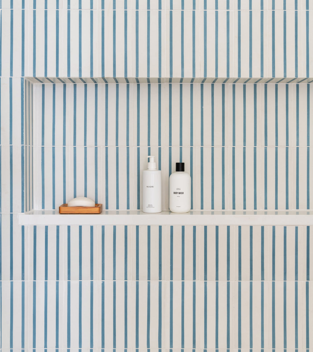 A shower niche lined with High Line Hyannis 4x8 cement tiles featuring vertical blue stripes, holding a bar of soap on a wooden dish and two white bottles.