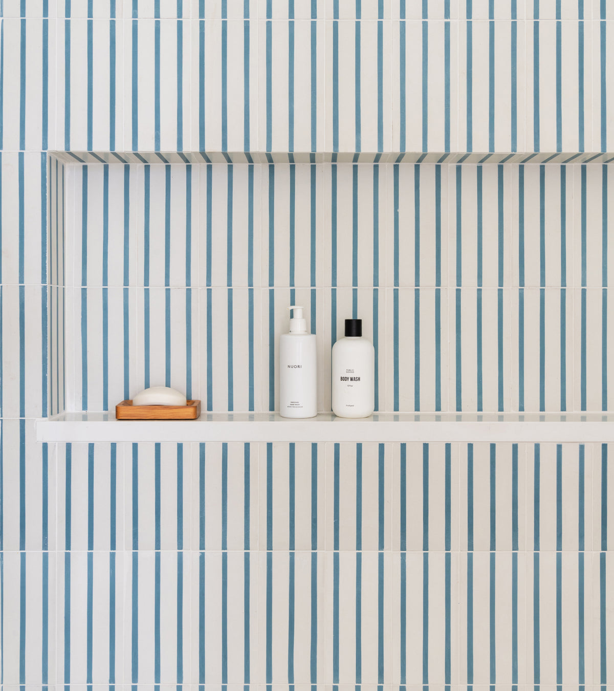 A shower niche lined with High Line Hyannis 4x8 cement tiles featuring vertical blue stripes, holding a bar of soap on a wooden dish and two white bottles.