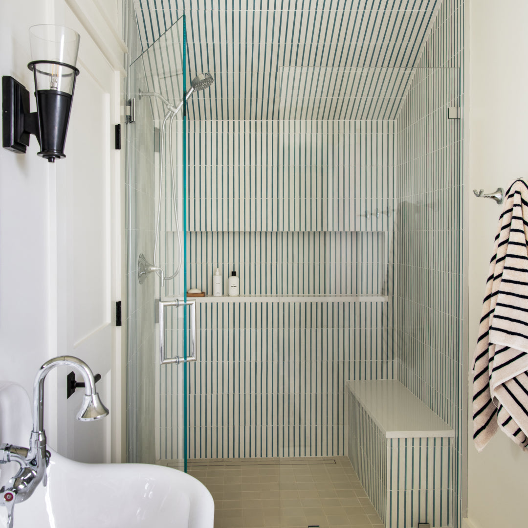 A shower stall lined with High Line Hyannis 4x8 cement tiles in thin blue and white vertical stripes, featuring a glass door, a built-in bench, and a recessed storage shelf.