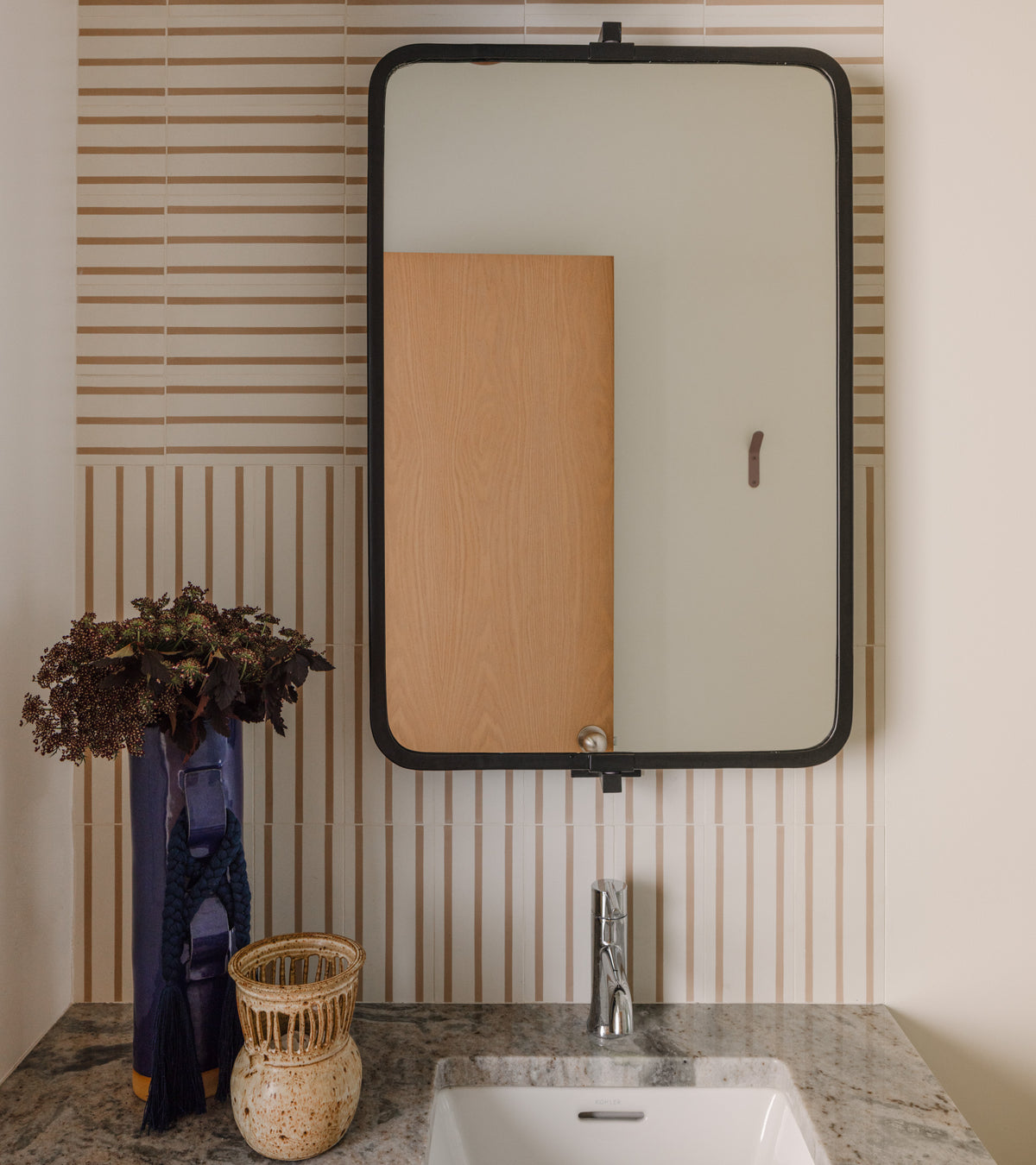A modern bathroom vanity features a rectangular mirror, marble countertop, potted plant in a blue vase, and woven basket. The wall showcases Zia Tile's High Line Jaipur Pink 4x8 tiles. A wooden door is visible in the mirror's reflection.