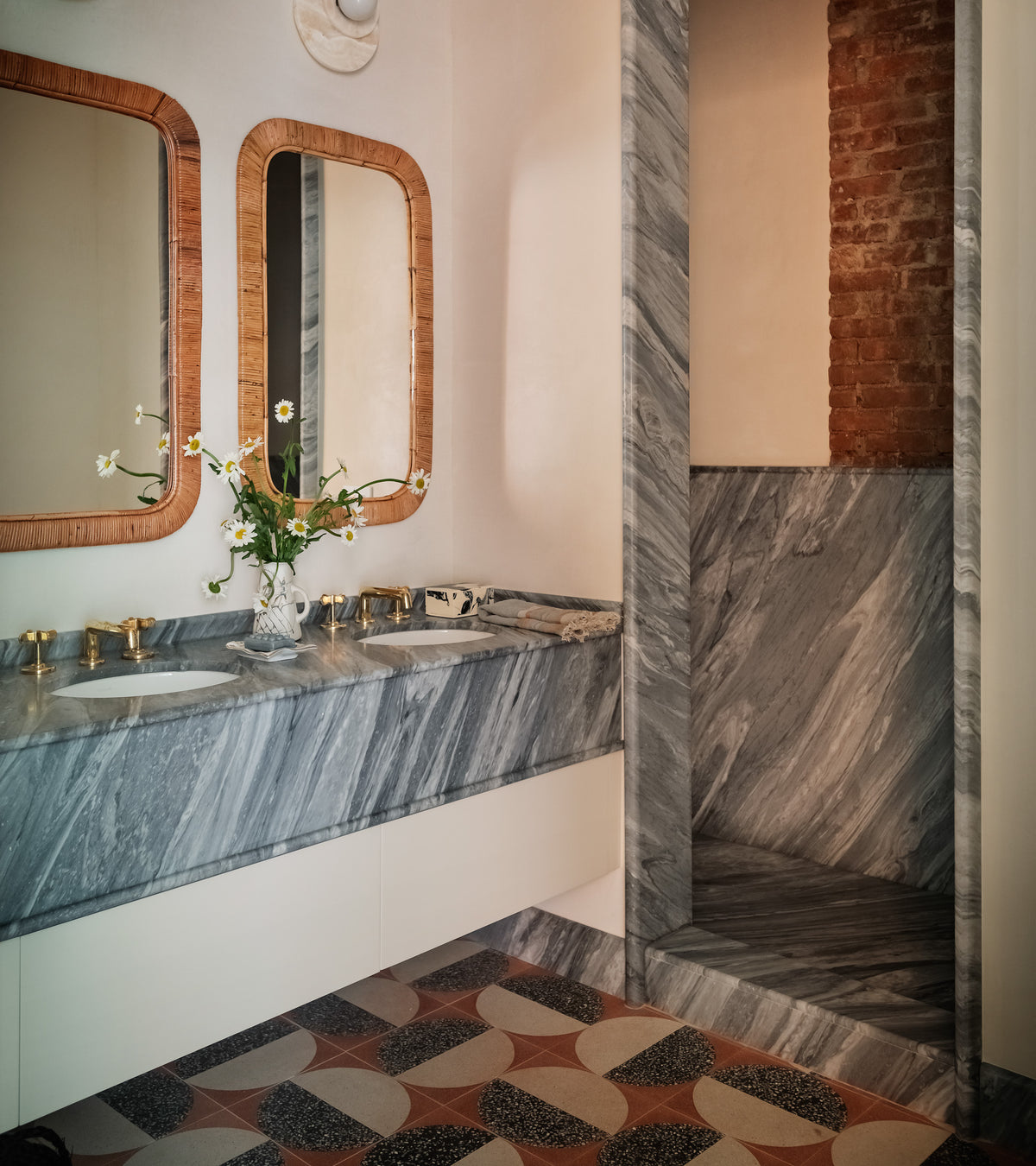 A stylish bathroom featuring marble countertops, gold faucets, dual rectangular mirrors, an open marble shower, exposed brick, a floral vase, and modern Highball Rust 12x12 terrazzo cement tile flooring by Zia Tile.