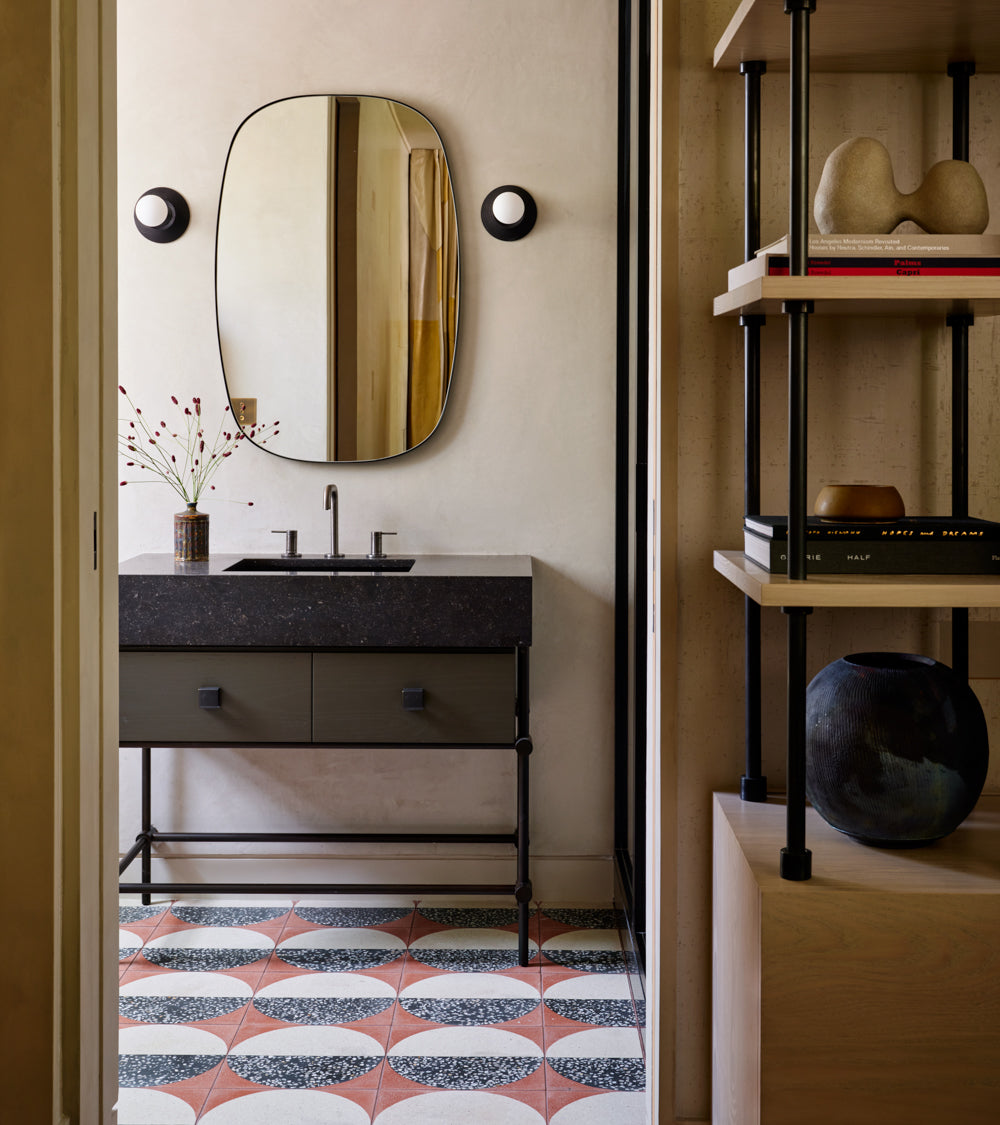 A modern bathroom features a black vanity, round mirror, wall sconces, and Zia Tile's Highball Rust 12x12 cement tile flooring. Open shelves hold decor and books, while a vase with branches rests on the vanity.