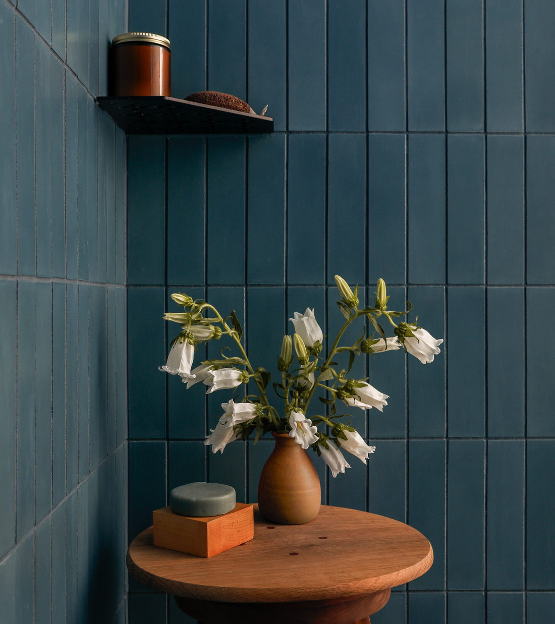 A small round wooden table with a brown vase of white flowers and soap on a tray sits before a vendor-unknown Hyannis 2x8 subway tile backsplash. A black shelf above holds a brown jar and sponge, adding to the kitchen scene.