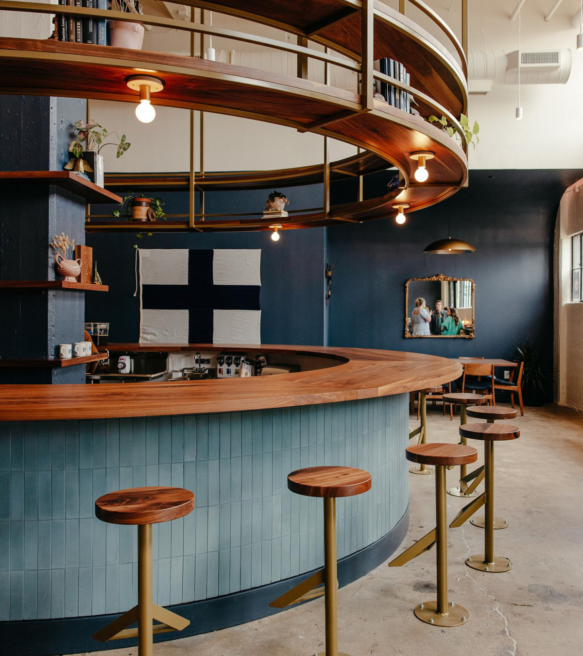 A modern café with a curved wooden bar, brass stools, circular shelves, and a blue-and-white flag. The bar front features Hyannis 2x8 tile by vendor-unknown. Warm lighting and a cozy seating area appear in the background.
