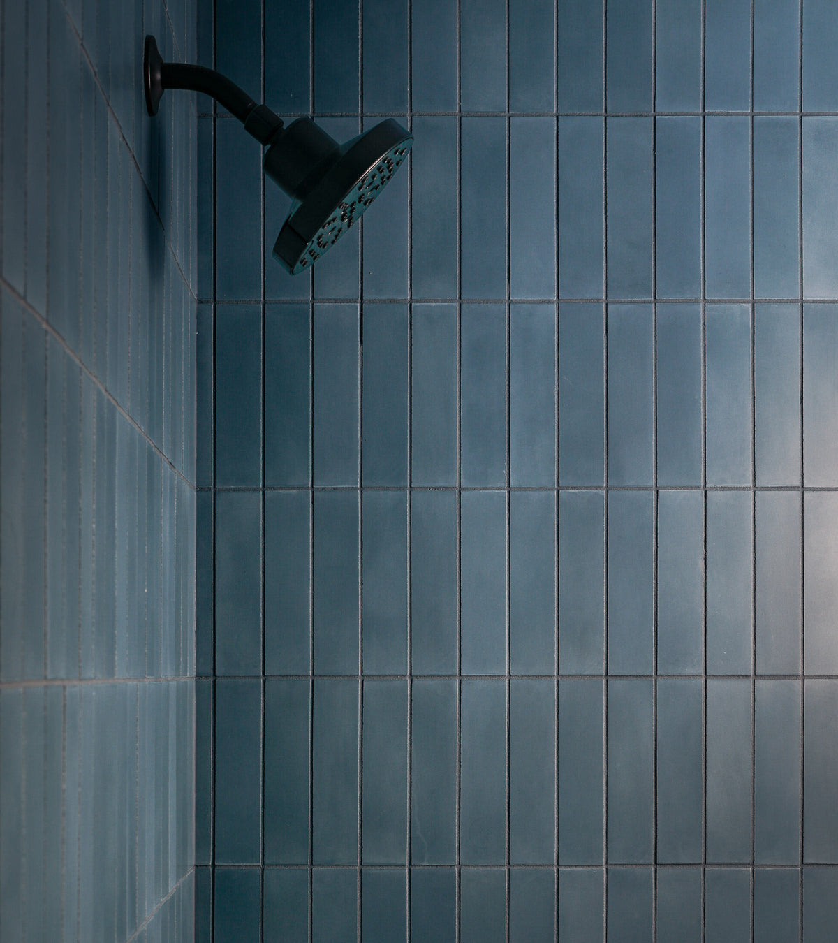 A matte black showerhead is mounted on a wall clad in vendor-unknown Hyannis 2x8 subway tiles arranged vertically, creating a modern, minimalist vibe in the shower.