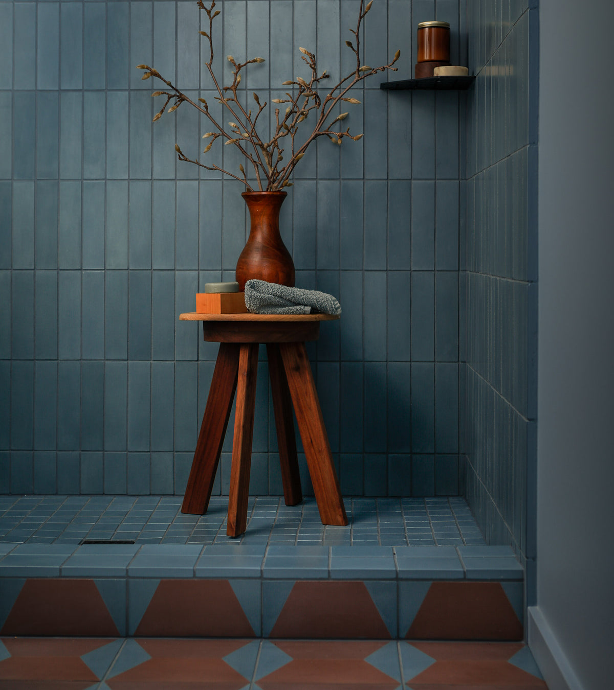 A modern bathroom features Zia Tile's Colorama Tyrian 8x8 blue handmade tiles on the floor and walls, with a wooden stool holding a brown vase of branches, folded towel, book, and a nearby wall shelf with a candle.