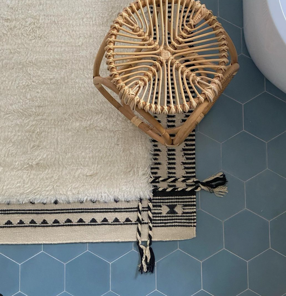 A Hyannis Hex tile floor by vendor-unknown in blue, paired with a black and cream patterned rug, a cream shaggy rug, and a round wicker stool near the edge of a white bathtub.