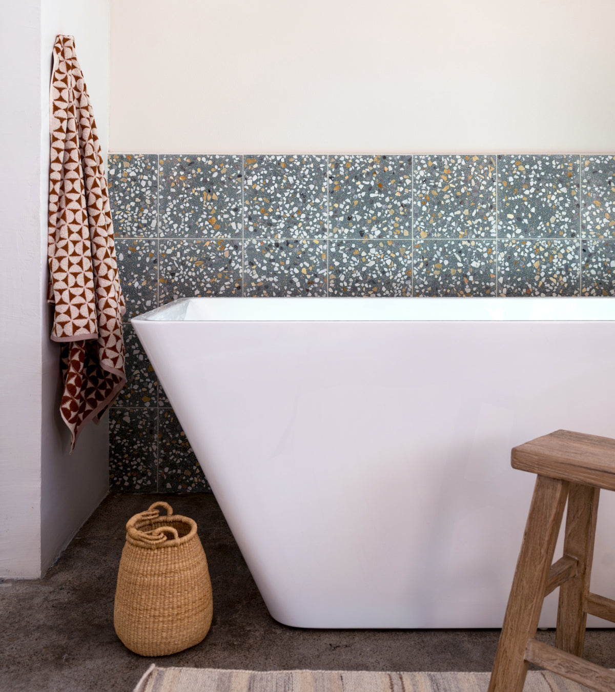 A modern bathroom features a white freestanding bathtub, Idyllwild 12x12 by Zia Tile in blue and gray Terrazzo on the wall, a geometric-patterned towel, wicker basket, and wooden stool on a textured rug.