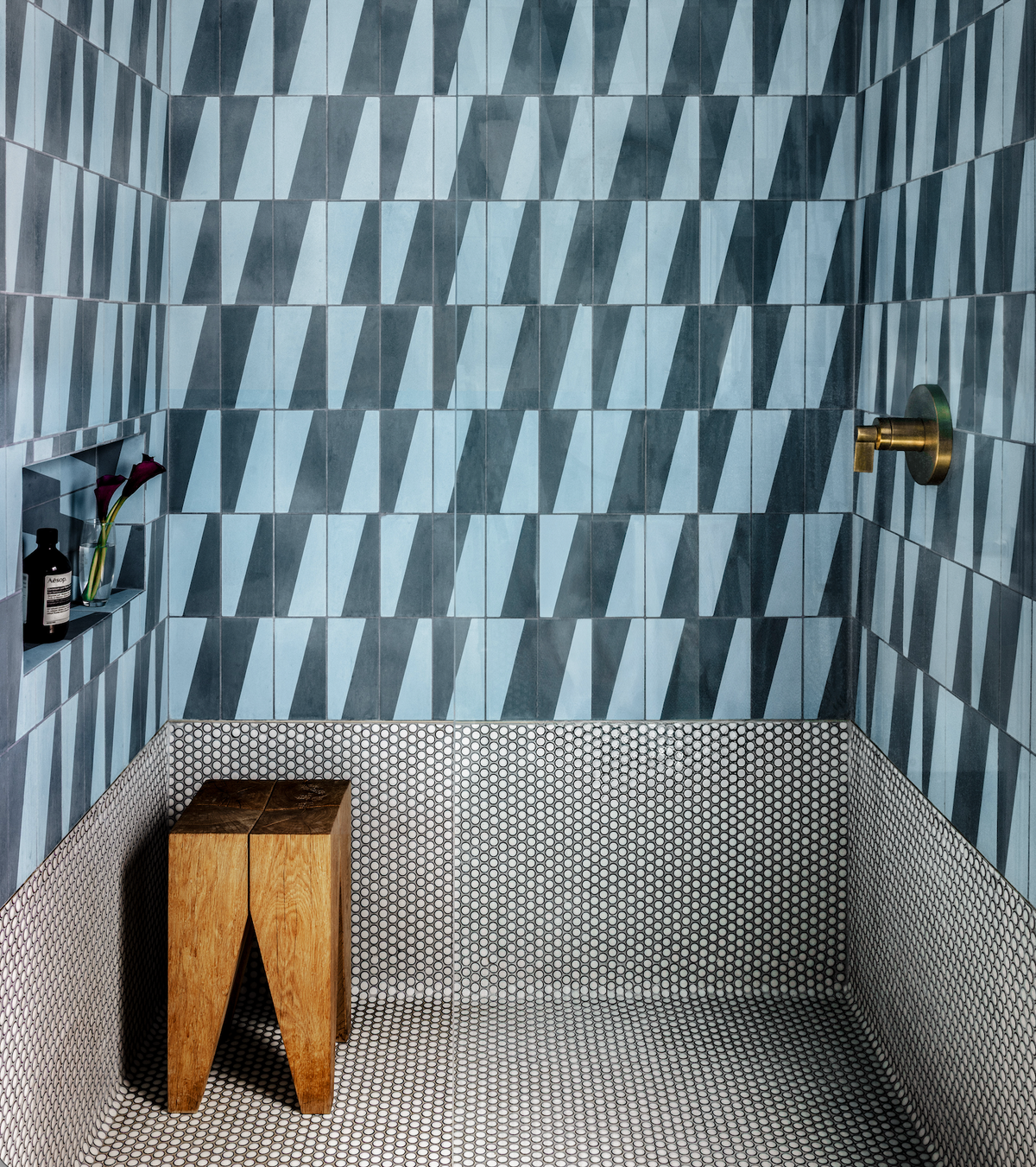 A shower featuring Zia Tile's Incline Hyannis 4x8 blue geometric wall tiles, small round mosaic floor tiles, a brass fixture, recessed shelf with toiletries and a flower, plus a wooden stool in the corner.