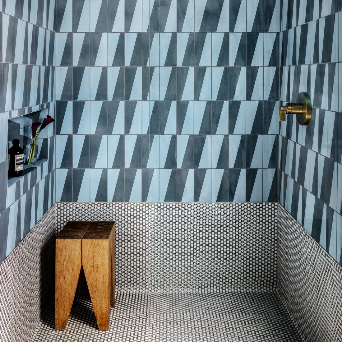 A shower with blue and gray geometric wall tiles, small round black-and-white floor tiles, a wooden stool, a brass shower fixture, and a built-in niche holding a bottle and flowers.