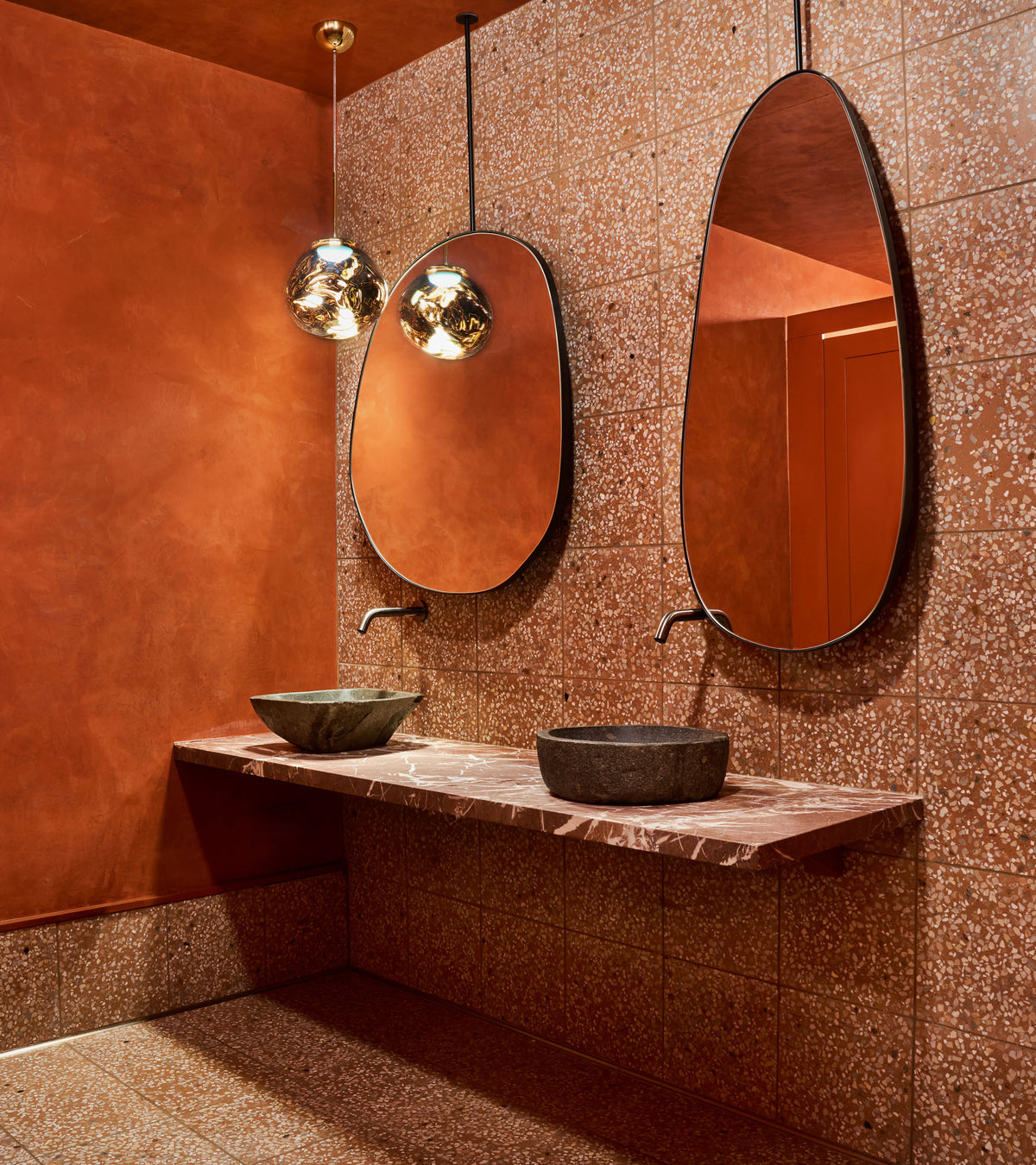 A modern bathroom featuring Indio 12x12 tiles from Zia Tile on the floor, terracotta walls, two irregular oval mirrors above a marble countertop with stone vessel sinks, and metallic pendant lights hanging from the ceiling.