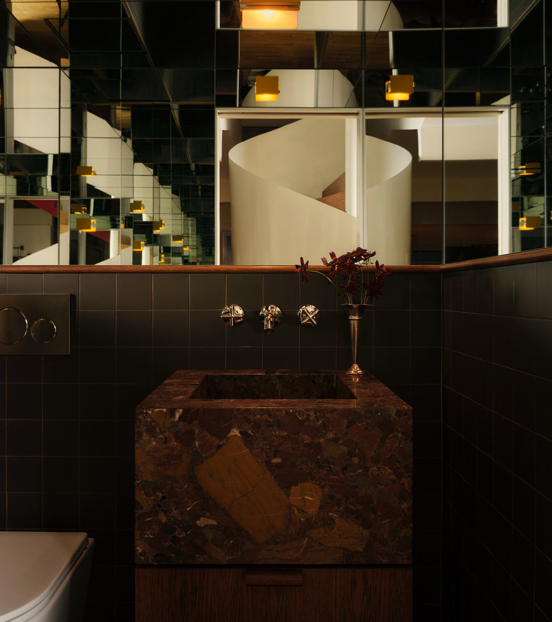 A modern bathroom featuring Ironwood 4x4 ceramic tiles by Zia Tile on the walls, a stone sink, wall-mounted faucets, and a vase with red flowers. The mirrored wall reflects a white spiral staircase and glowing yellow lights.