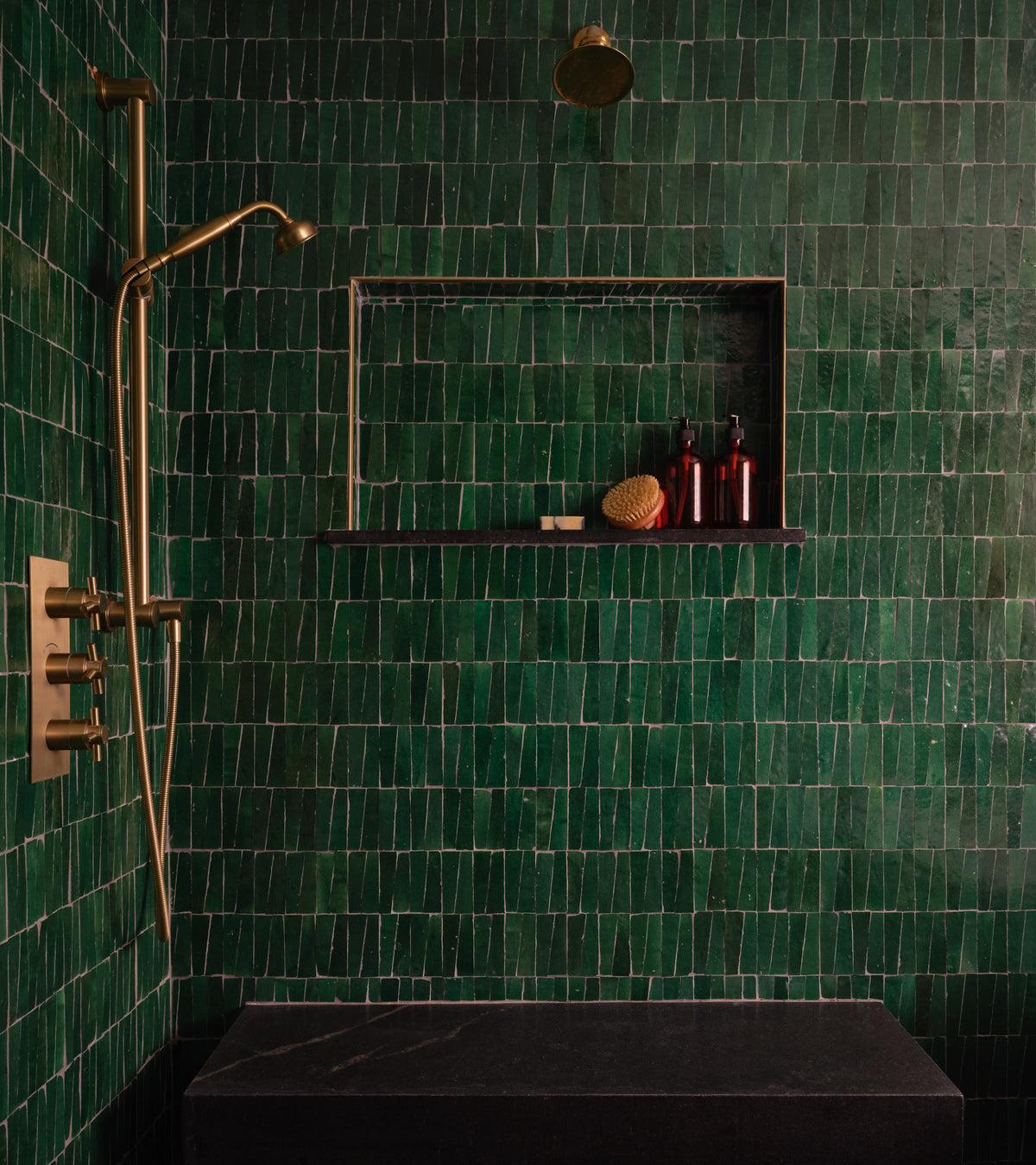 A modern shower features Zia Tile’s Jade 2x2 dark green zellige tiles, brass fixtures, and a built-in black shelf with red bottles and a sponge for a stylish, minimal look.