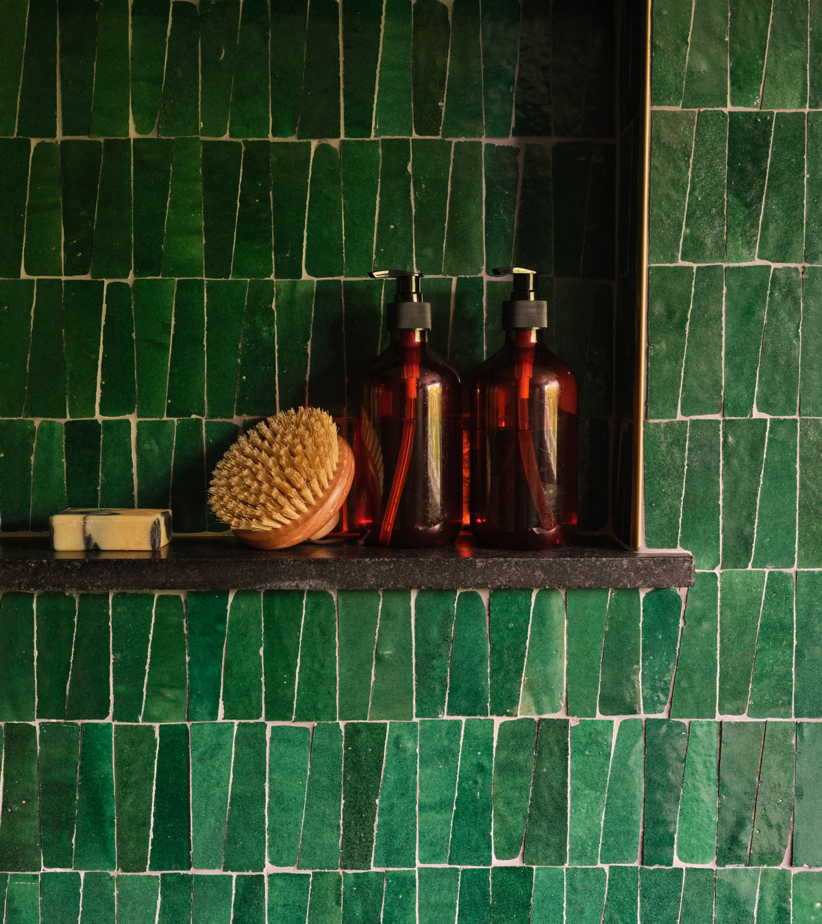 A shelf against a wall of glossy Jade 2x2 tiles by Zia Tile holds two amber pump bottles, a wooden bath brush with firm bristles, and a bar of soap.
