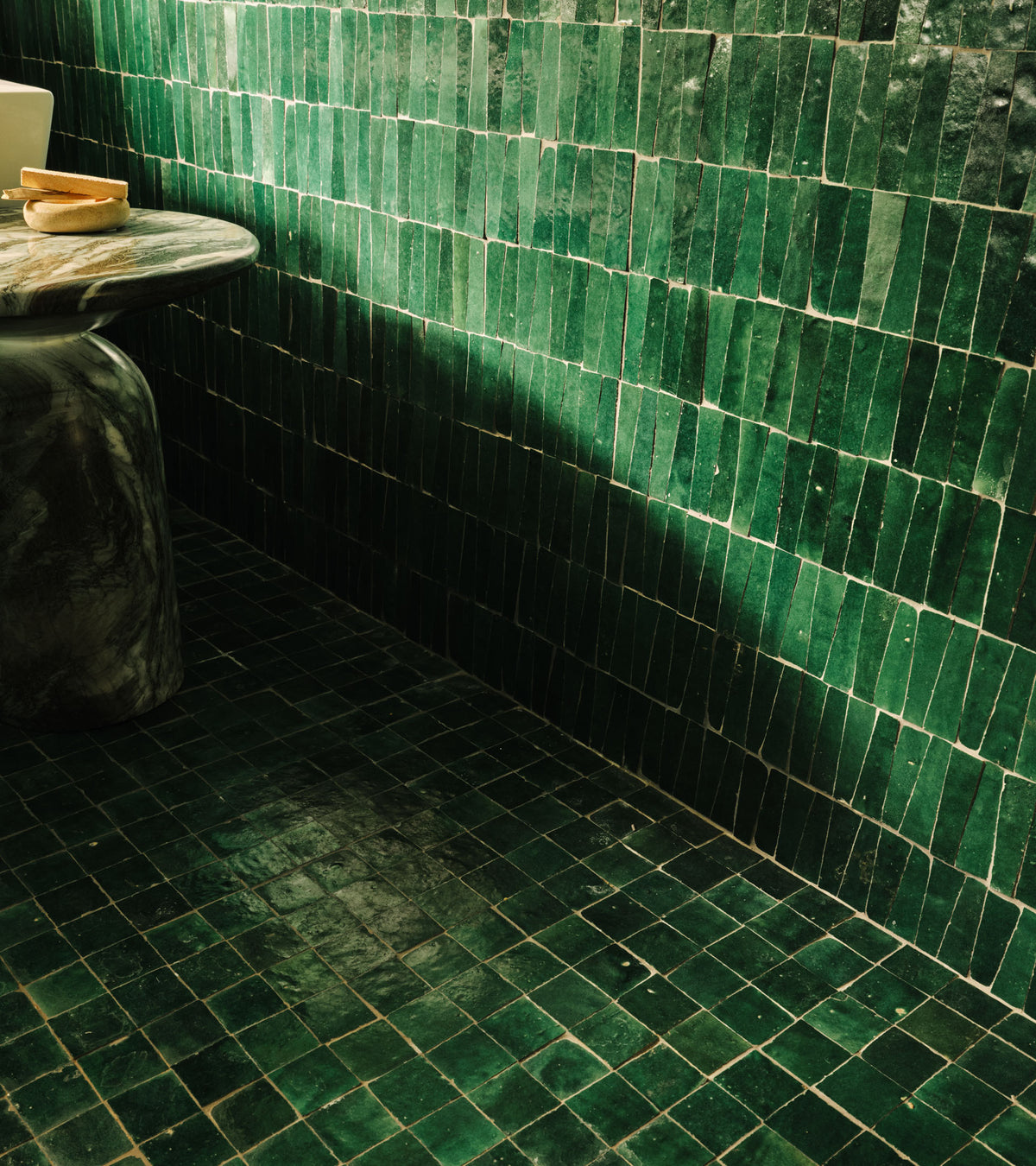 A bathroom corner features Zia Tile's Jade 2x2 tiles on the wall and floor, paired with a marble round pedestal table holding a small dish and bowl.