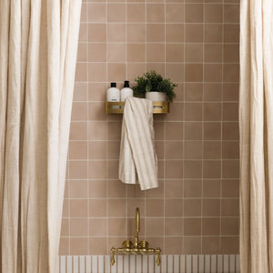 A beige-tiled bathroom wall with a brass faucet, cream shower curtains, a gold shelf, and striped towel—accented by vendor-unknown’s Jaipur Pink 4x4 tile for a subtle pop of color.