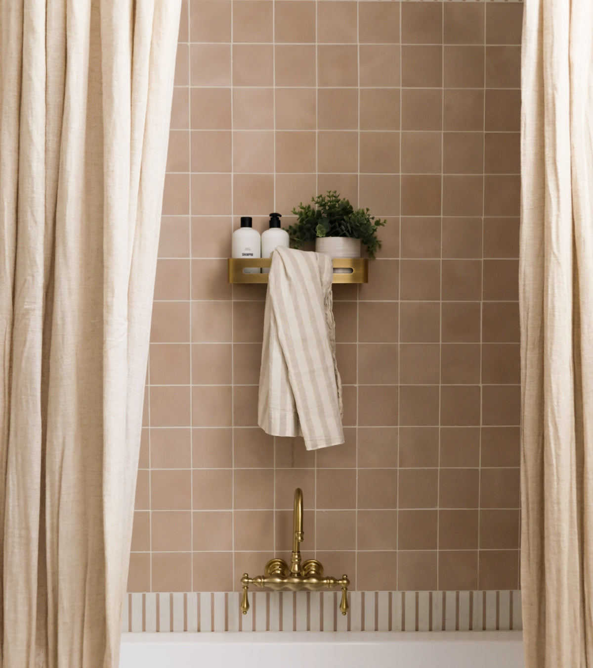 A beige-tiled bathroom wall with a brass faucet, cream shower curtains, a gold shelf, and striped towel—accented by vendor-unknown’s Jaipur Pink 4x4 tile for a subtle pop of color.