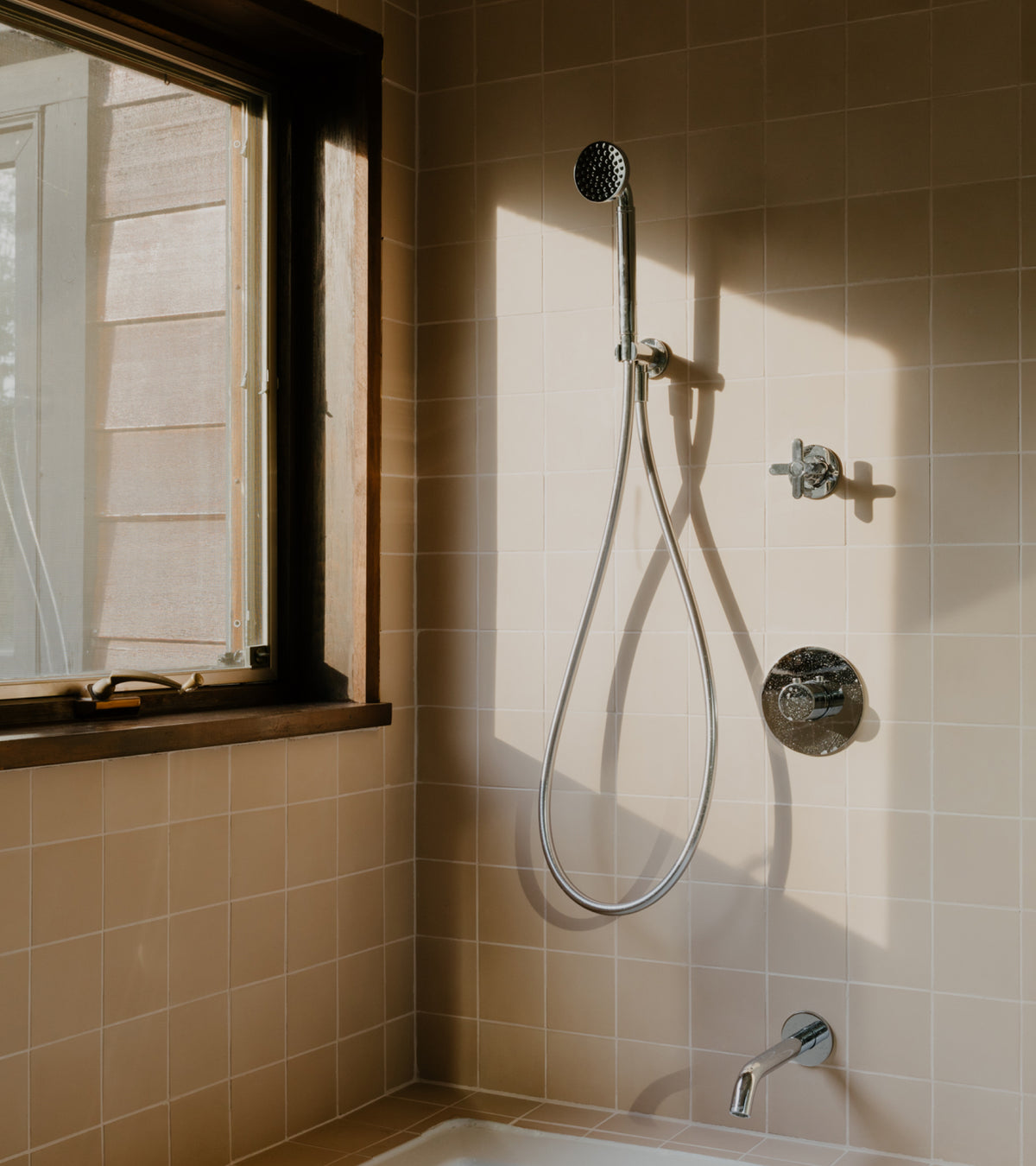 A wall-mounted shower head and handheld sprayer set installed against a Jaipur Pink 4x4 cement tile wall next to a window.
