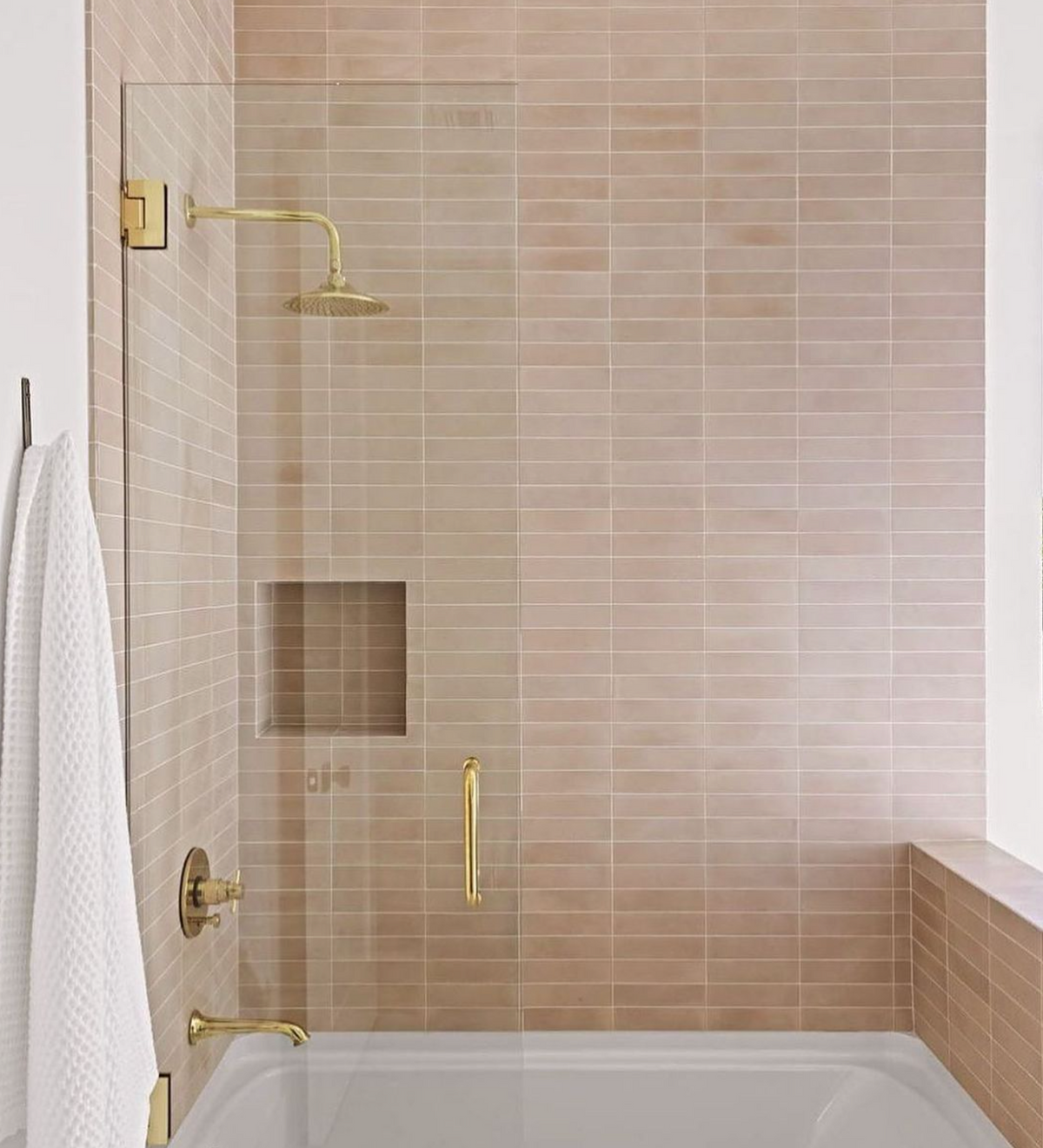 A modern bathroom with beige subway tiles, a glass shower door, gold fixtures, a built-in niche, and a white towel by the tub is accented with elegant handmade Jaipur Pink 2x8 tiles from vendor-unknown.
