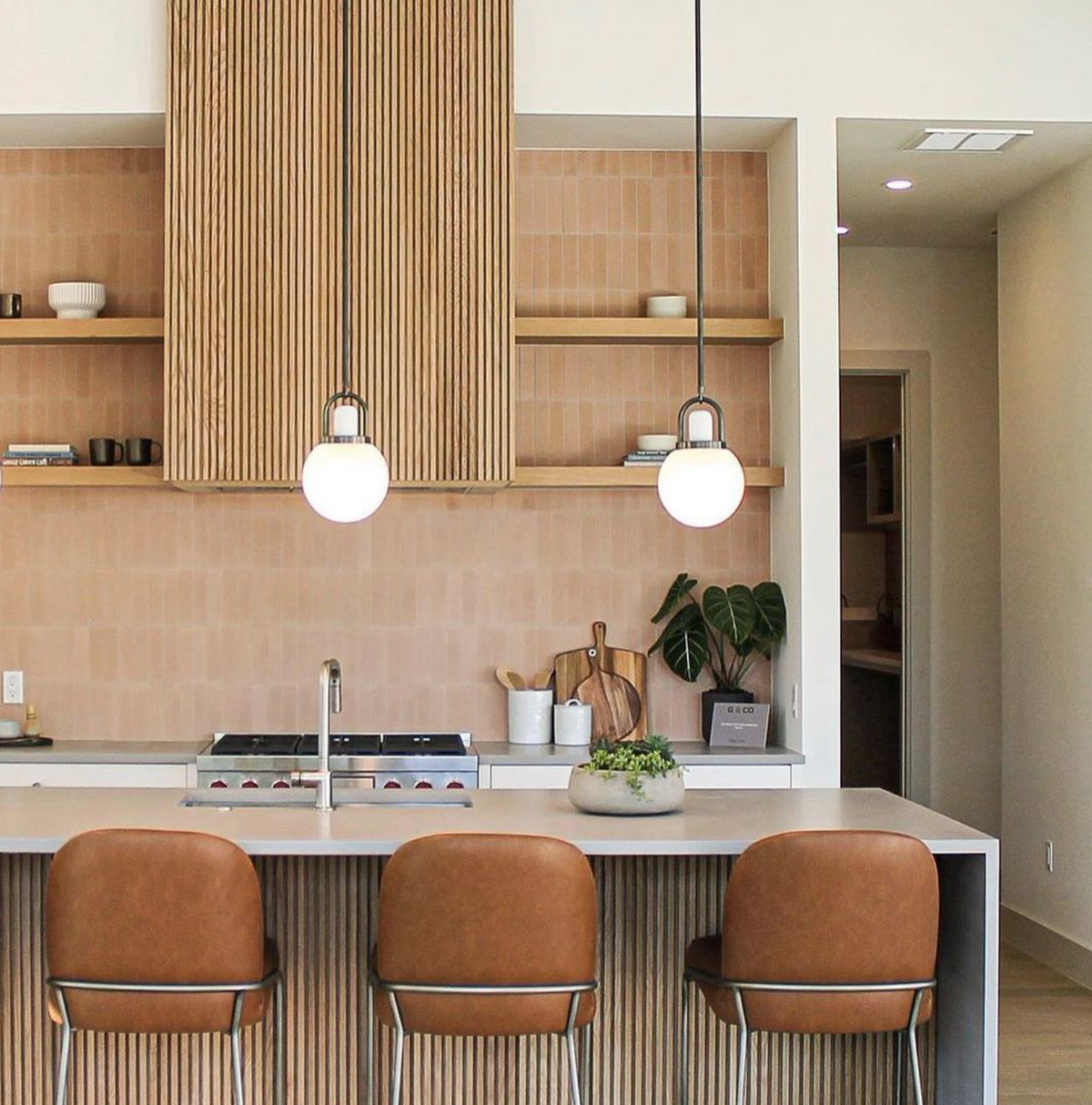 Modern kitchen with light brown cabinets, open wooden shelves, and a Jaipur Pink 2x8 backsplash. Three brown leather barstools sit at a white island lit by pendant lights. Plants and kitchen items decorate the counter and shelves.
