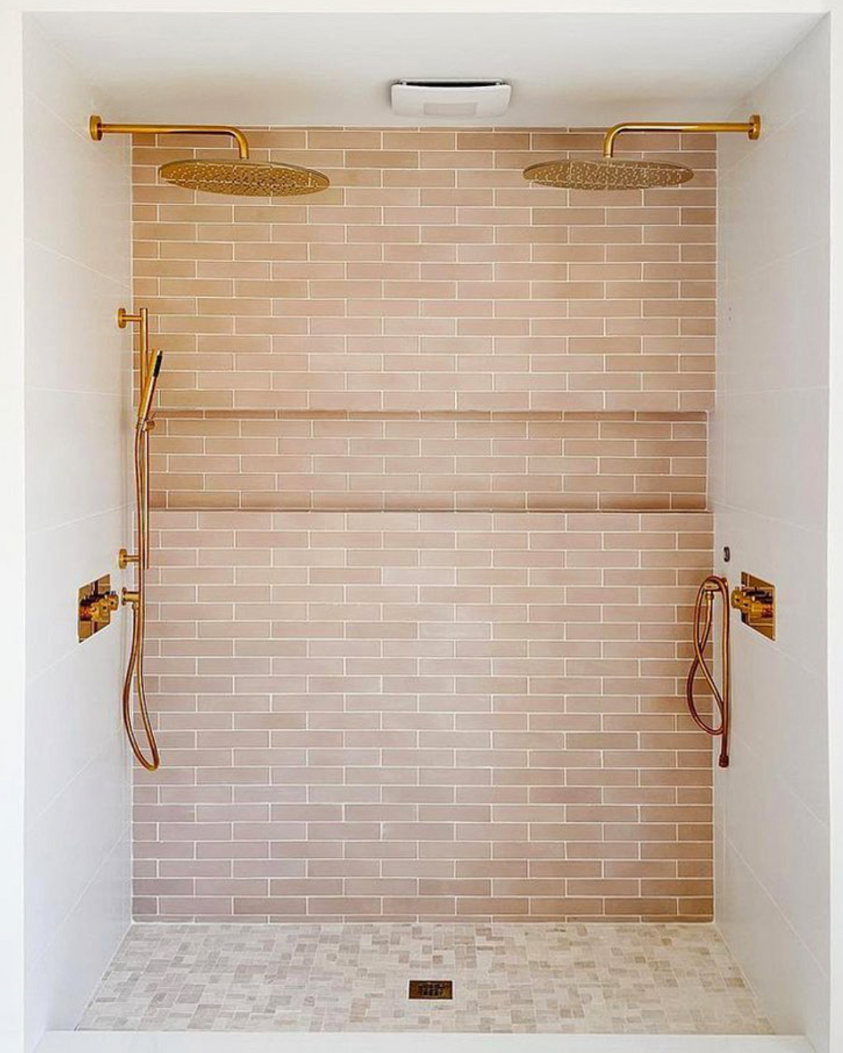 A bright, minimalist shower features Jaipur Pink 2x8 tiles from vendor-unknown, twin gold rainfall showerheads, matching gold fixtures, a built-in shelf, and a light mosaic cement tile floor.