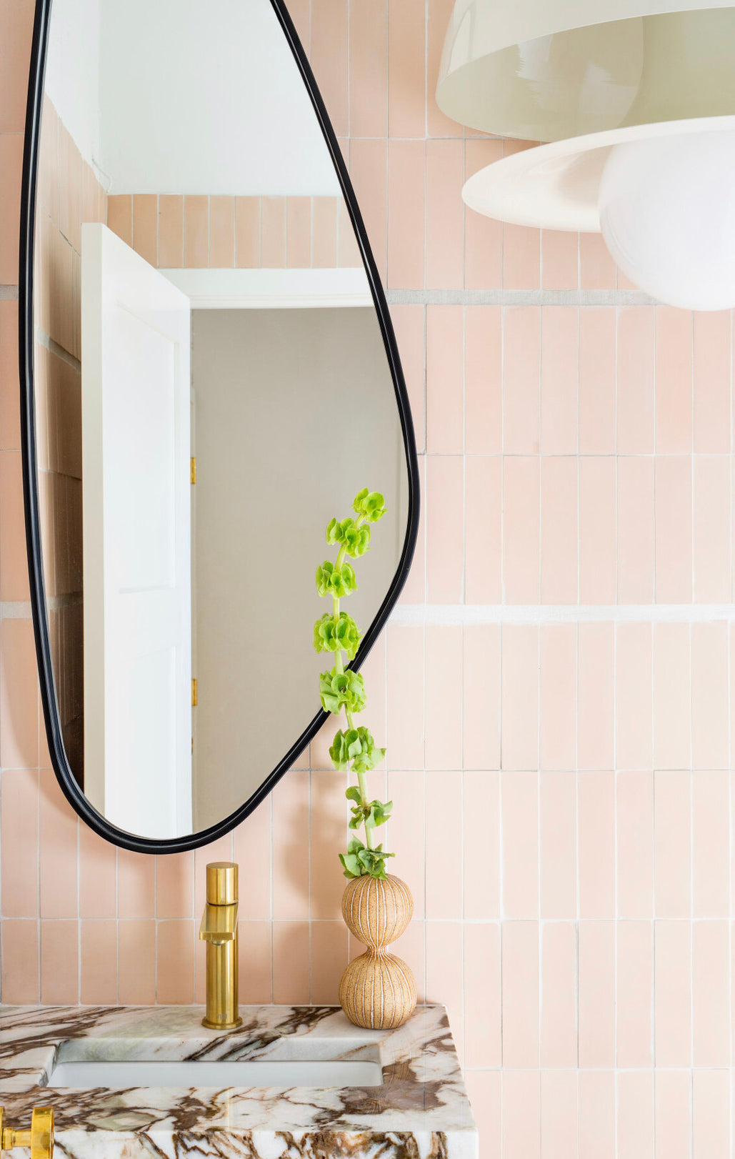 A modern bathroom features Jaipur Pink 2x8 handmade tile walls (vendor-unknown), an irregular mirror, marble countertop with gold faucet, beige door, and a tall green plant in a textured beige vase.