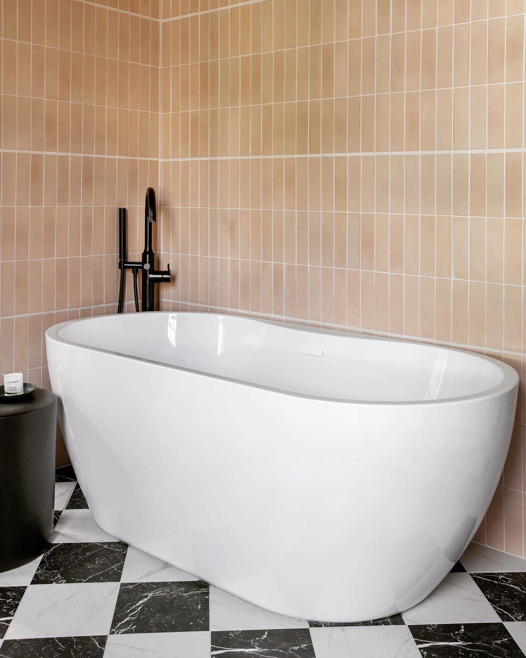A modern bathroom with a white freestanding bathtub, Jaipur Pink 2x8 tiles by vendor-unknown on the wall, black and white checkered cement floor tiles, a black side table with a candle, and a sleek black faucet next to the tub.