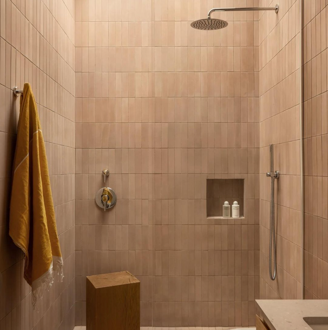 Minimalist walk-in shower with Jaipur Pink 2x8 tiles by vendor-unknown, ceiling-mounted rain shower, built-in niche for two bottles, wooden stool, yellow towel on the wall, and sleek handheld shower fixture.