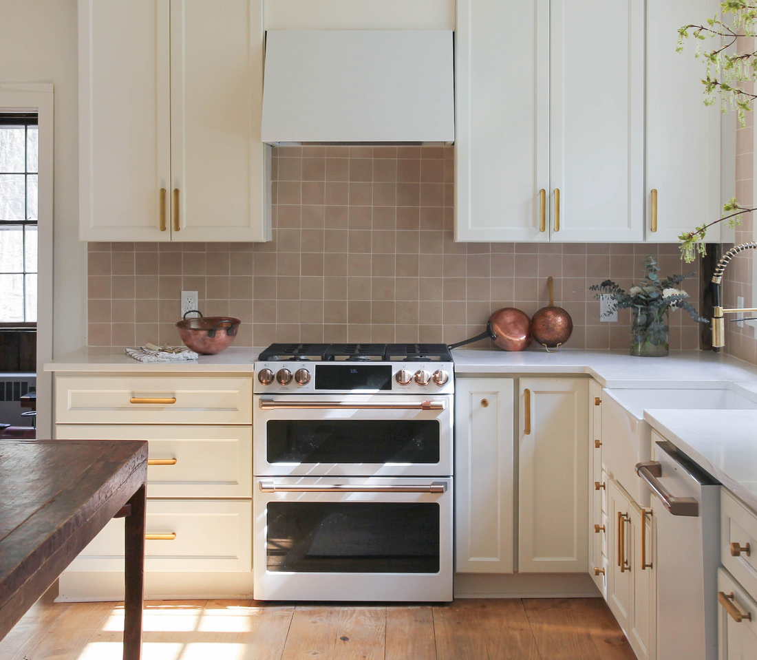 A bright, modern kitchen features cream cabinets with gold handles, a stainless steel oven, and a Jaipur Pink 4x4 tile backsplash by vendor-unknown above white countertops. Copper bowls, greenery, wood floors, and a large window give a warm touch.