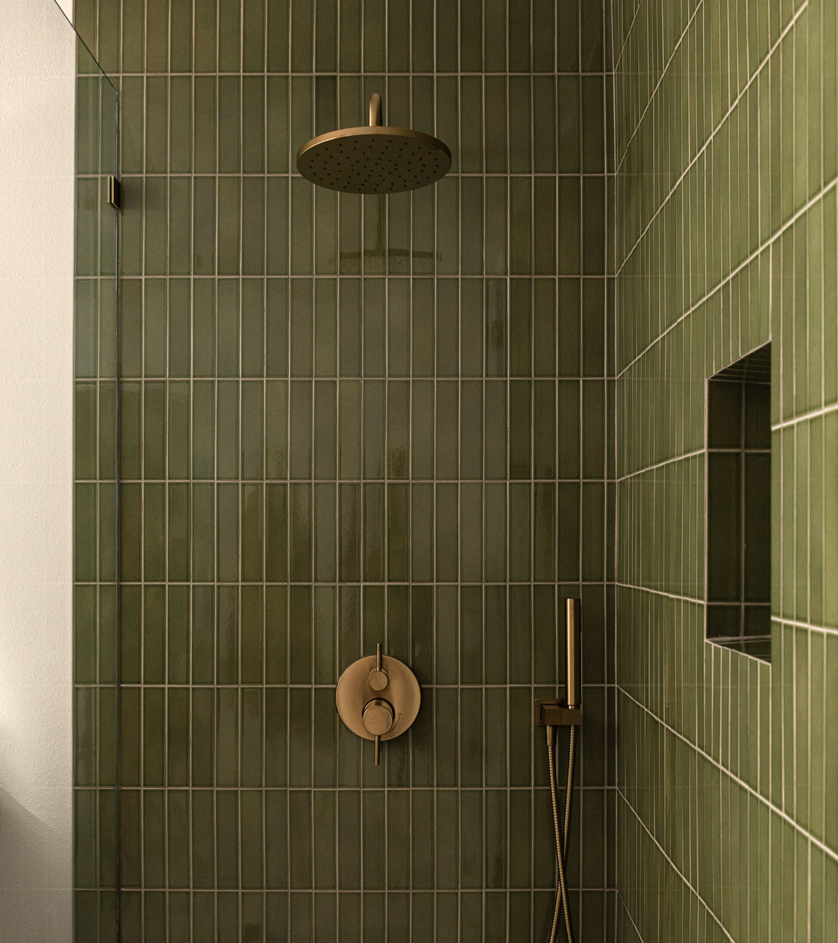 A modern shower featuring Zia Tile’s Kelp Forest 2x8 glossy green ceramic tiles, a round overhead shower head, built-in wall niche, and gold shower controls with handheld wand.