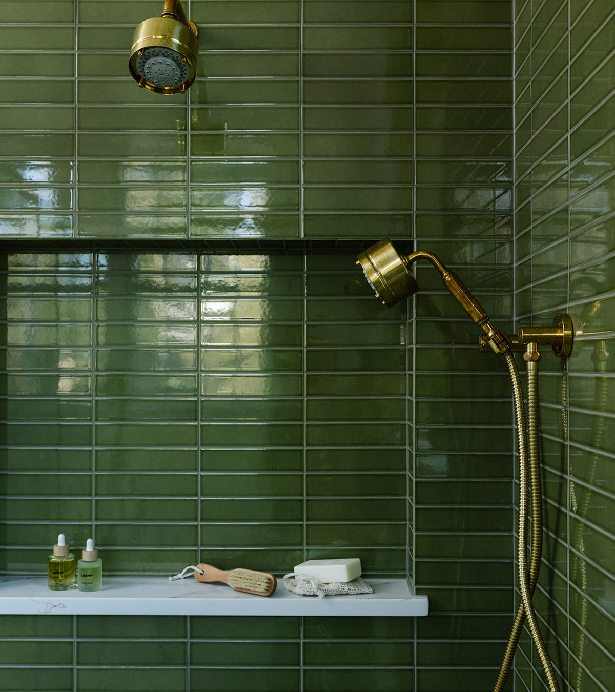 A shower features Zia Tile’s Kelp Forest 2x8 architectural-grade glossy olive green ceramic tile, a gold showerhead with matching handheld sprayer, and a white shelf for toiletries. Light reflections accentuate the tile’s vibrant sheen.