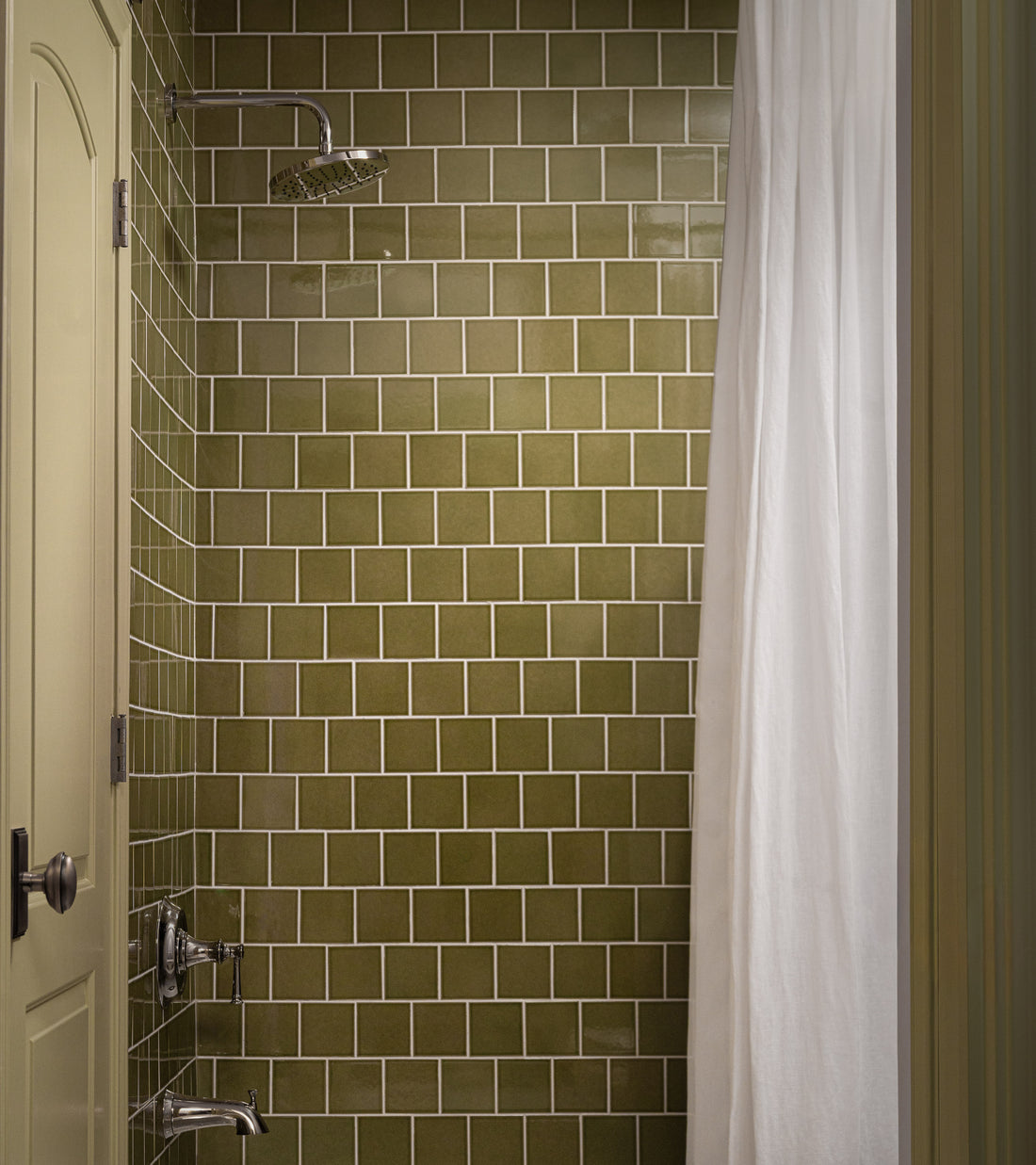 A shower features Zia Tile’s Kelp Forest 4x4 olive green glossy ceramic tile, a silver showerhead with two handles, and a partially drawn white curtain. On the left, there’s a beige door with a dark handle.