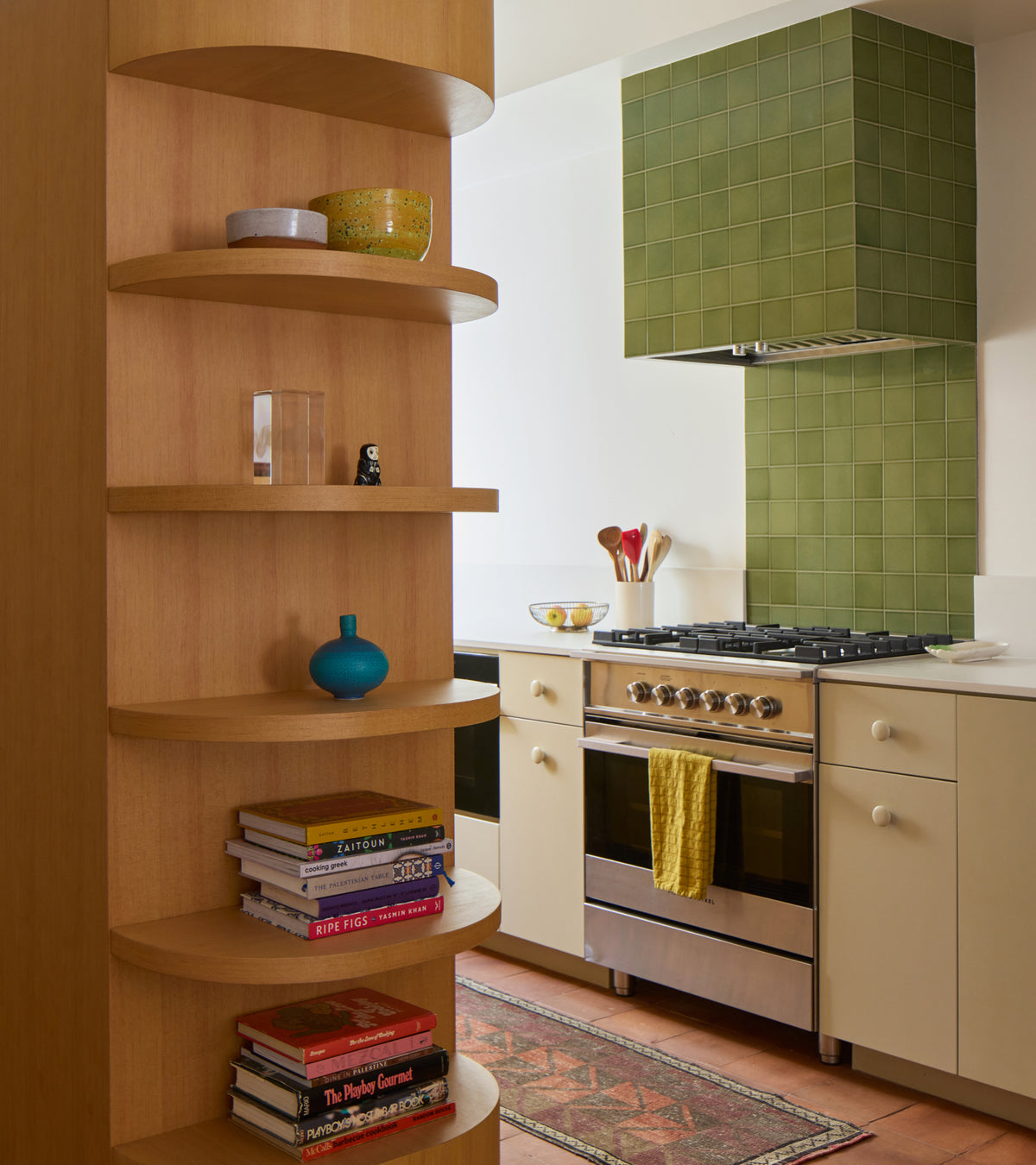 A kitchen featuring a stainless steel stove with a glossy Kelp Forest 4x4 ceramic tile backsplash and a curved wooden shelving unit stocked with books and decor.