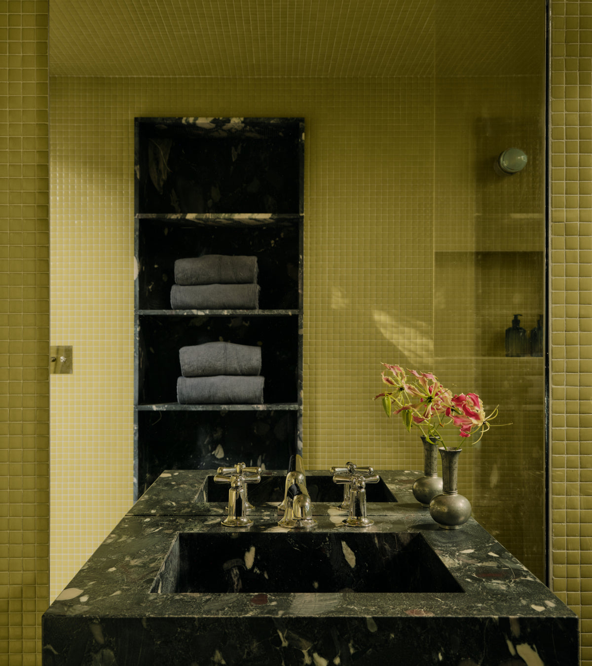 A dark stone bathroom vanity with a double sink and silver faucets, set against walls covered in Kiwi 1x1 olive-green square tiles by Zia Tile.