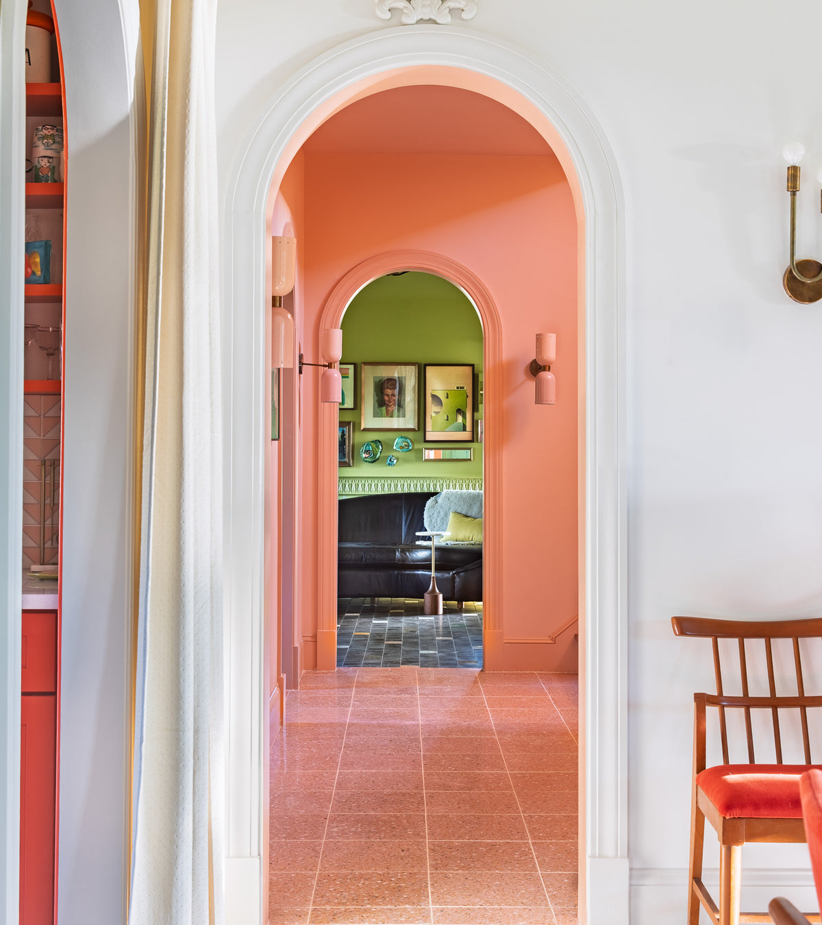 A bright hallway with peach walls and arched doorways has Las Palmas 12x12 cement tiles from Zia Tile, leading to a green room with art, a black couch, a white chair, and a wooden chair with an orange cushion to the right.