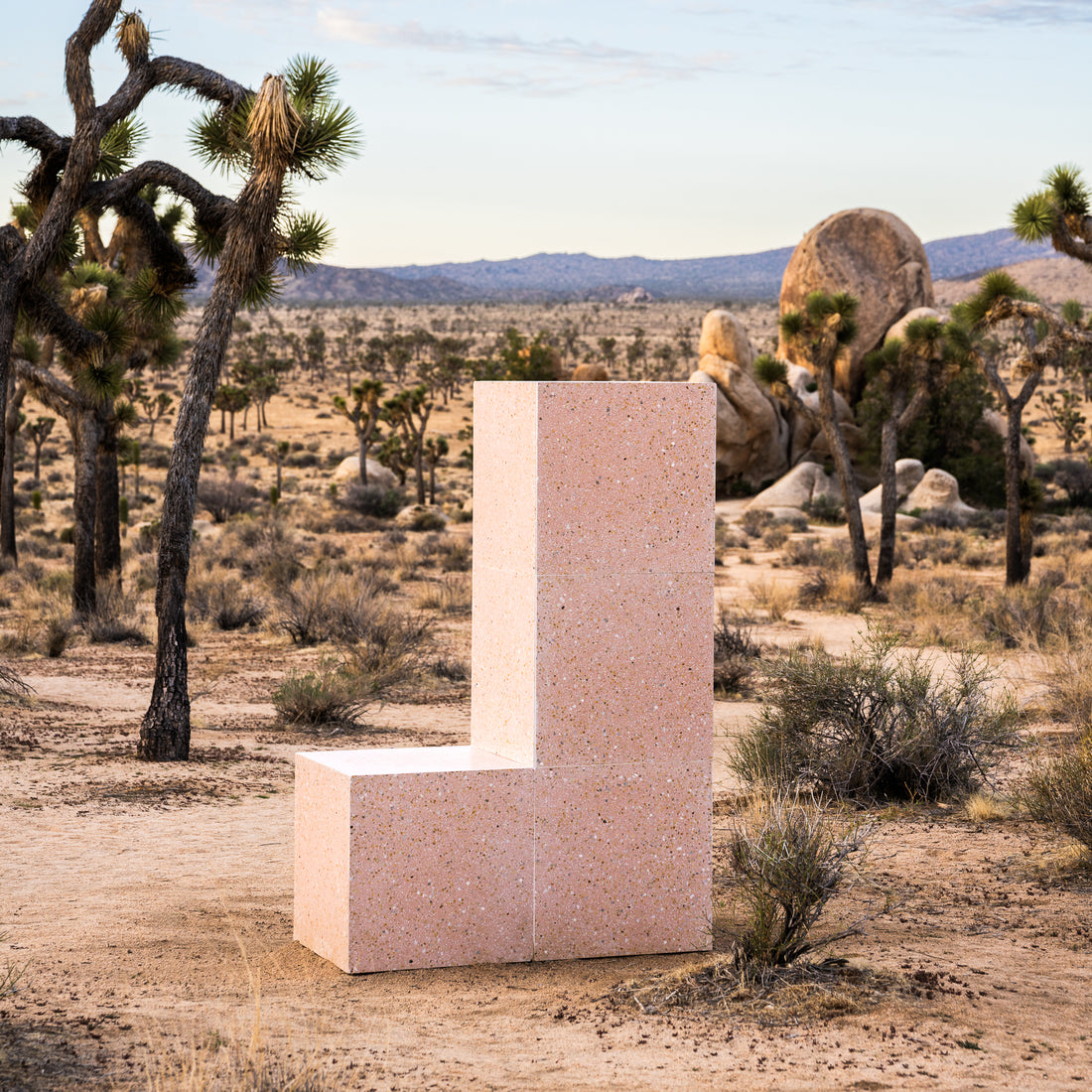 The Las Palmas 24x24 chair by Zia Tile features a geometric L-shape in light pink terrazzo, set in a desert landscape with Joshua trees and sparse bushes, reflecting the artisanal charm of handmade tile under a partly cloudy sky.