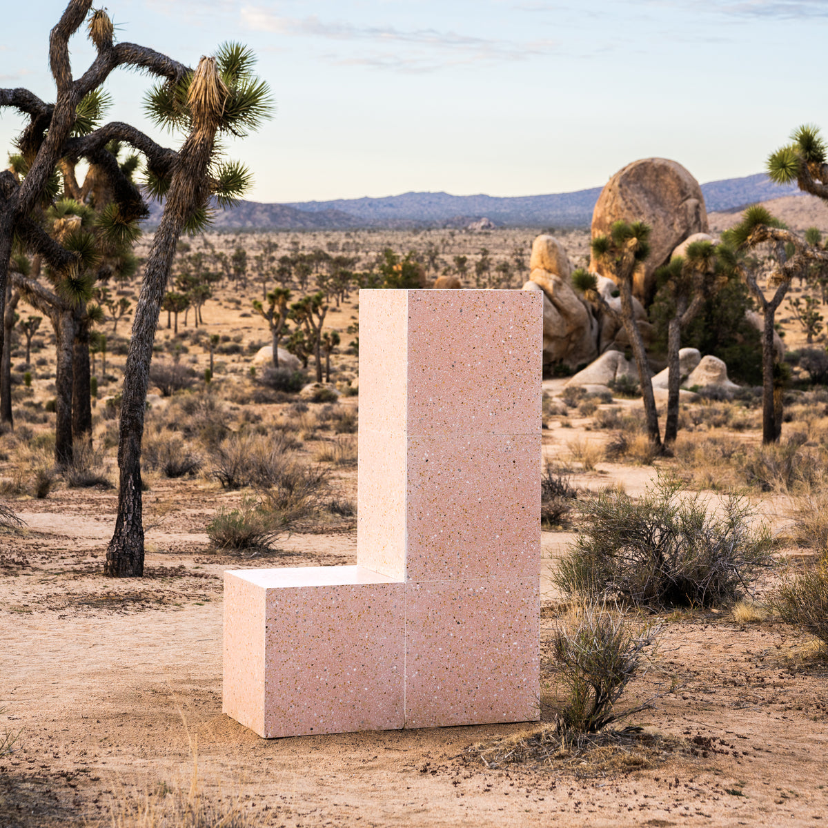 The Las Palmas 24x24 chair by Zia Tile features a geometric L-shape in light pink terrazzo, set in a desert landscape with Joshua trees and sparse bushes, reflecting the artisanal charm of handmade tile under a partly cloudy sky.