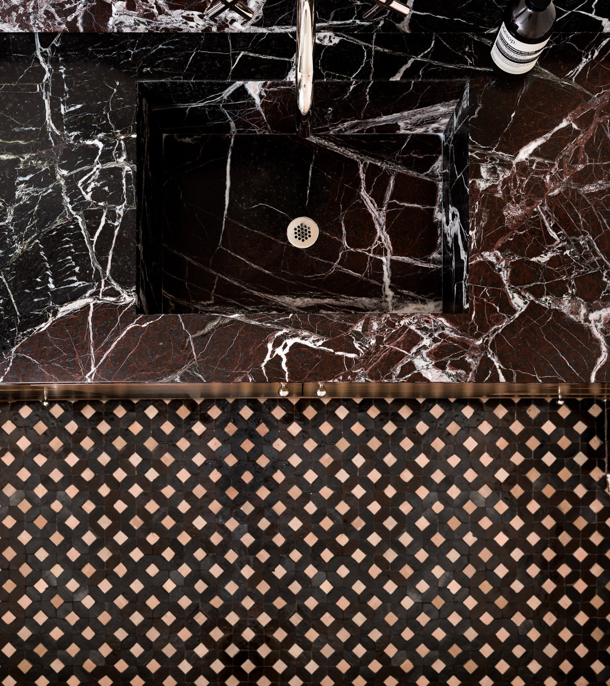 A black marble sink with white veining sits below a silver faucet, atop a matching countertop. The bathroom backsplash features Zia Tile's Lattice 6—a geometric pattern in black, brown, and beige Moroccan tile.