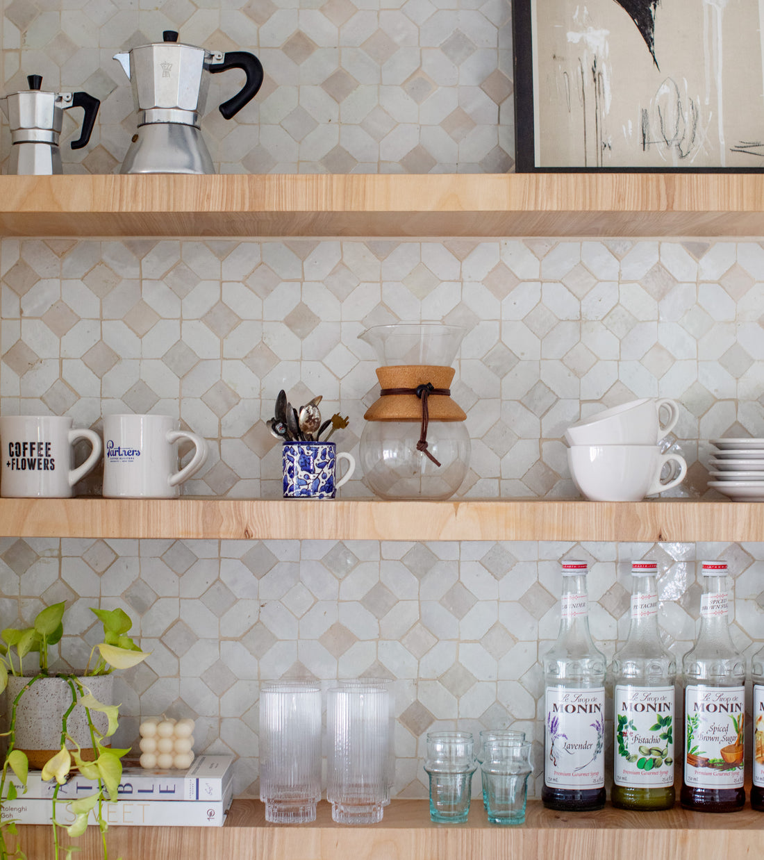A wooden kitchen shelf displays mugs, bowls, cups, glasses, and more against a Zia Tile Lattice 4 Moroccan tile backsplash.