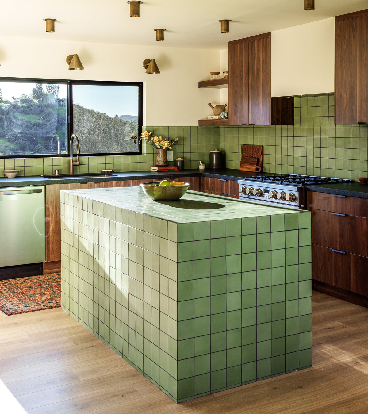 A modern kitchen features Leaf 4x4 green square backsplash tile by vendor-unknown on countertops and a large island, complemented by wooden cabinets, stainless steel appliances, open shelves, and a window with a view of greenery.