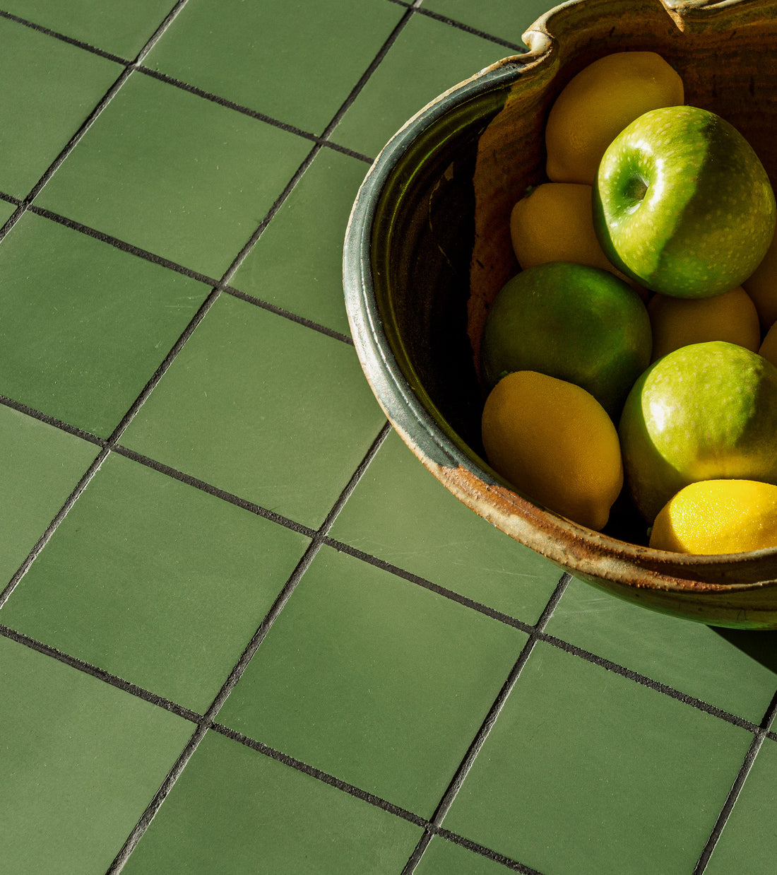 A ceramic bowl with green apples and yellow lemons sits on vendor-unknown’s Leaf 4x4 green tile, with sunlight casting gentle shadows across the scene.