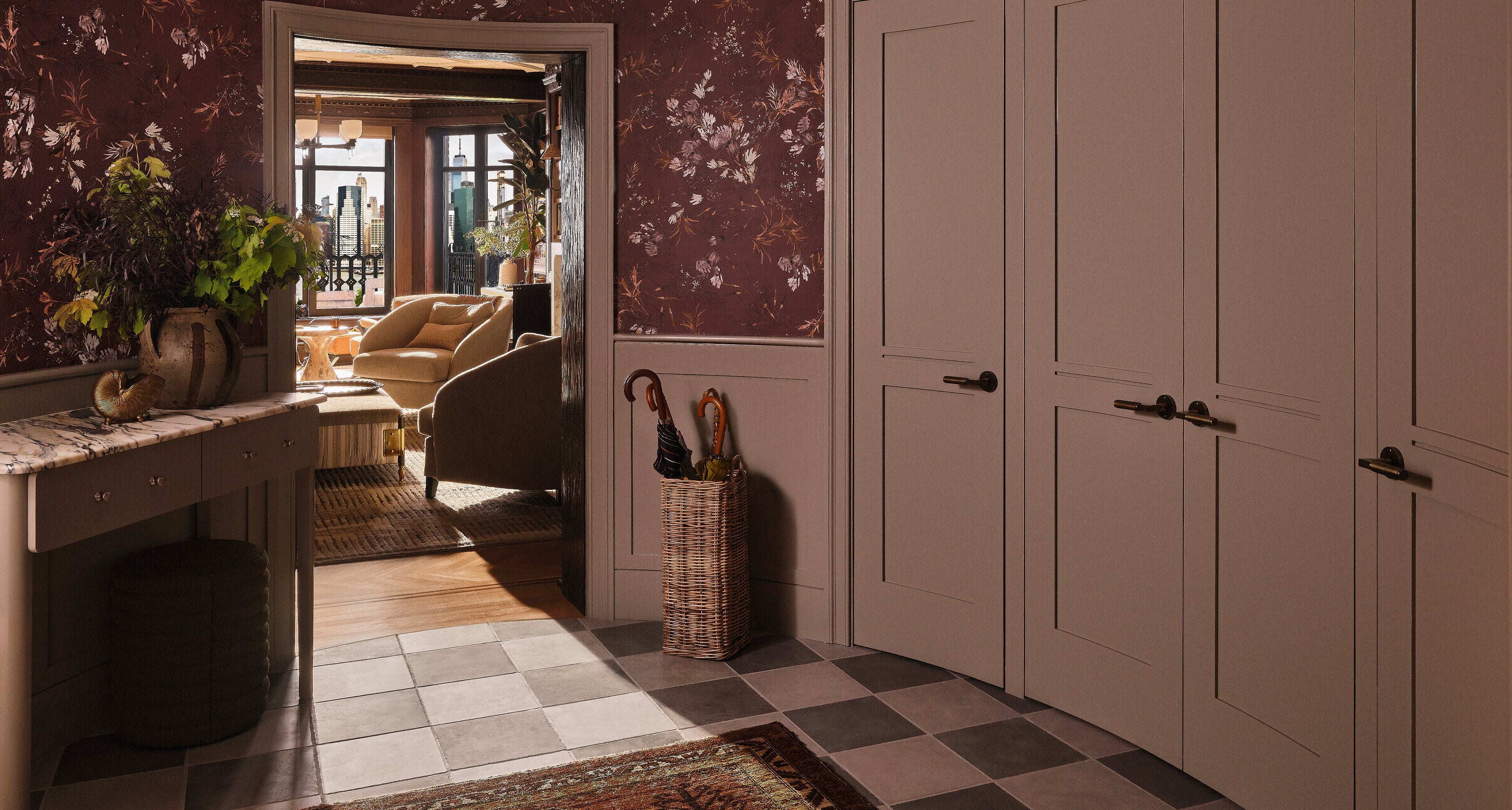 An elegant entryway with floral wallpaper, checkered tile floor, built-in cabinets, a wicker umbrella stand, and a marble console. An open door reveals a warmly lit room with armchairs and large windows.